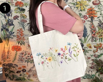 Könnte beinhalten: Eine weiße Canvas-Tragetasche mit einem Blumendesign, das verschiedene Blumen und Blätter in den Farben Rosa, Lila, Blau und Grün zeigt. Die Tasche hat einen langen Riemen und wird von einer Person getragen.