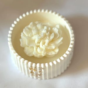May include: A white ceramic candle holder with a raised, textured rim and gold accents. A white wax flower is centred in the holder.