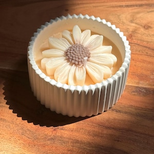 May include: A white, ridged candle holder with a daisy-shaped candle. The candle has a light beige flower with a brown center. The candle sits in a light orange wax. The candle holder is on a wooden surface.