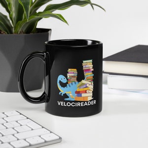 May include: Black ceramic mug featuring a blue dinosaur reading a stack of books. The mug has the text "VELOCIREADER" printed on the bottom.