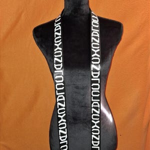 Könnte beinhalten: Schwarze Schärpe mit weißem Text, der vertikal angezeigt wird. Der Text scheint ein sich wiederholendes Buchstabenmuster zu sein. Die Schärpe ist über einer schwarzen Schaufensterpuppe vor einem orangefarbenen Hintergrund drapiert.