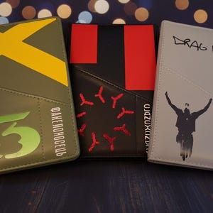 Peut inclure: Trois porte-passeports rectangulaires aux designs distincts. Celui de gauche est vert olive avec un X jaune et du texte vert. Le central est noir avec des accents rectangulaires rouges et des coutures rouges. Celui de droite est gris avec le texte DRAG PATH.