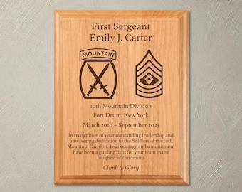 May include: A wooden plaque with a brown stain, engraved with the text "First Sergeant Emily J. Carter", "10th Mountain Division", "Fort Drum, New York", "March 2010 - September 2023", and "Climb to Glory". The plaque also features a military insignia with crossed swords and a chevron.