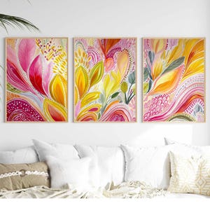 May include: Three framed abstract watercolor paintings with floral designs. The artwork features vibrant colors, including pink, yellow, and orange, with intricate details. The paintings are displayed above a white couch with pillows.