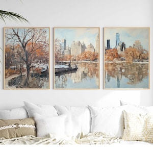 Op de afbeelding: Een set van drie ingelijste prints die een stadsbeeld met een meer op de voorgrond tonen. De prints zijn in een gedempte kleurenpalet van bruinen, grijzen en blauwen. De stadsgezichten worden weerspiegeld in het meer, waardoor een symmetrische compositie ontstaat.