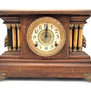 May include: Antique wooden mantel clock with a round clock face featuring Roman numerals. The clock has decorative columns, ornate brass accents, and a detailed design on the wooden base. The clock is a warm brown color.