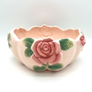 May include: A light pink ceramic bowl with a scalloped rim, adorned with raised rose and leaf details. The bowl features a large pink rose in the center and smaller roses on the sides, with green leaves. A decorative floral accent.