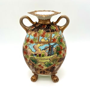 May include: A decorative ceramic vase with a landscape scene featuring a windmill, trees, and a blue sky. The vase has two handles, a square top, and three feet. The color palette includes brown, gold, red, and blue.
