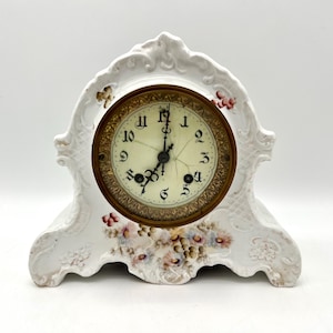 May include: White porcelain mantel clock with a floral design. The clock face is gold-toned with black numerals and hands. The clock has a decorative, ornate design with floral accents in pink, red, and yellow.