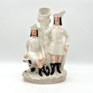 May include: A vintage ceramic figurine depicting two figures in white outfits with black boots and hats, standing next to a tree-like structure. A dog is seated near one figure. The base reads "ROBIN HOOD".