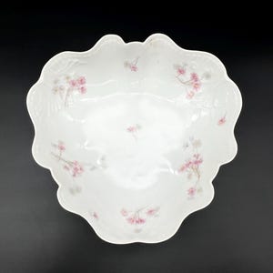 May include: A white, scalloped-edge serving bowl with delicate pink floral patterns. The bowl has a shallow, rounded shape and is decorated with clusters of pink flowers and green leaves. The floral design is repeated around the rim and in the center of the bowl.