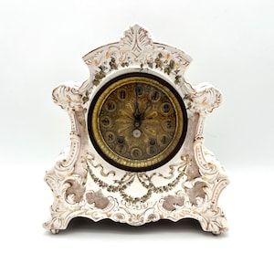 May include: Antique white mantel clock with ornate detailing. The clock features a gold-toned face with black numerals and hands. The clock's frame is decorated with floral and ribbon motifs, adding to its vintage charm.