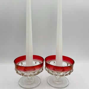 May include: Two glass candle holders with a red rim and clear base, each holding a tall white candle. The candle holders have a decorative design around the rim and a round base. The background is plain white.