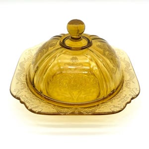 May include: Amber-colored glass butter dish with a domed lid and a square base. The lid has a round knob and is decorated with etched floral patterns. The base also features an etched design. The dish is made of transparent glass.