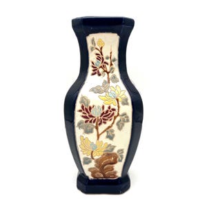 May include: A navy blue ceramic vase with a floral design. The vase has a cream-colored center panel with a detailed illustration of flowers in shades of yellow, burgundy, and brown. The vase is approximately 7 inches tall.