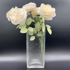 May include: A clear, textured glass vase holds three cream-colored roses with green leaves. The rectangular vase has a rough, bubbly surface. The roses are in full bloom, with multiple petals.