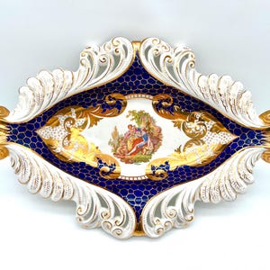 May include: An ornate, decorative porcelain tray with a white, gold, and navy blue design. The tray features a central oval scene with figures, surrounded by gold filigree and a navy blue honeycomb pattern. The edges are white with a sculpted leaf design.