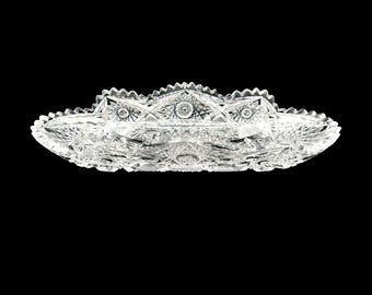 May include: A clear, oval-shaped crystal serving dish with intricate starburst and geometric patterns. The dish has a scalloped edge and is designed to catch and reflect light, creating a sparkling effect. Ideal for serving appetizers or desserts.