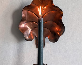 Puede incluir: Un candelero de cobre con un diseño en forma de hoja y una vela encendida. La vela es de color verde oscuro y el candelero tiene un borde festoneado.