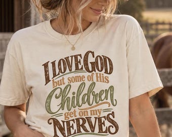 May include: A cream-colored short-sleeve t-shirt featuring the brown, orange, and green distressed text I LOVE GOD but some of His Children get on my NERVES printed on the front. The shirt has a crew neckline and a relaxed, casual fit.