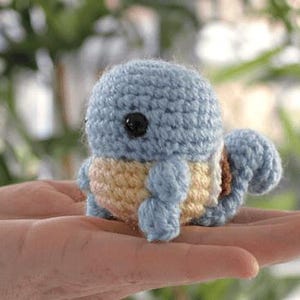 May include: A handmade crochet Squirtle plushie in light blue, yellow, and peach. The small, round figure has black button eyes and a curled tail. The plushie is held in a person's open hand.