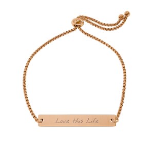 Peut inclure: Un bracelet réglable de couleur or rose composé d'une fine chaîne à maillons carrés et d'une barrette métallique rectangulaire. La barrette porte l'inscription gravée "Love this Life" et le bracelet dispose d'un fermoir coulissant.