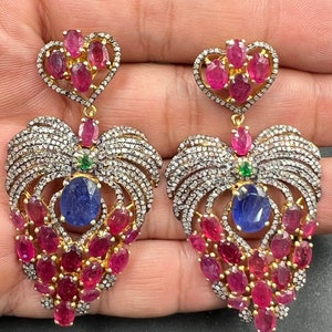 May include: A pair of dangle earrings with a heart-shaped design. The earrings are made of gold and feature a large blue gemstone surrounded by smaller pink gemstones and clear diamonds.