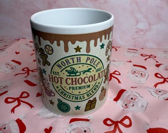 Könnte beinhalten: Keramikbecher mit weißem Innenraum und braunem Rand. Der Becher zeigt den Text "North Pole Hot Chocolate Premium Christmas Blend" und festliche Illustrationen. Der Becher steht auf einer rosa Oberfläche mit Weihnachtsmann- und Schleifenmustern.