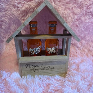 Könnte beinhalten: Zwei Aperol Spritz-Flaschen in einem holzförmigen Träger in Hausform. Die Flaschen sind orangefarben und tragen die Aufschrift "Apero Spritz". Der Träger trägt den Text "Petra 5 Aperol Bar".