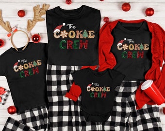 May include: A set of four black t-shirts with the text "The C*OKAE CREW" in gold glitter letters. The shirts are decorated with a red Santa hat, a gingerbread cookie, and a green Christmas tree. The shirts are worn by a family of four, with the adults wearing black and white plaid pajamas and the children wearing black and white checkered pajamas.