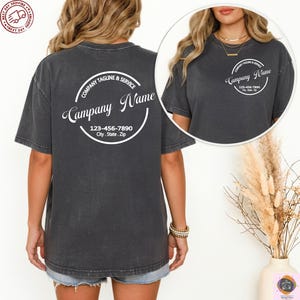 May include: Dark gray t-shirt with a circular design on the back that includes the text "Company Name", "Company Tagline & Service", and a phone number and address. The shirt is short-sleeved and has a relaxed fit.