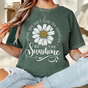 A woman wearing a green t-shirt with a white flower design and the text "When you can't find the sunshine, be the sunshine" printed on it.