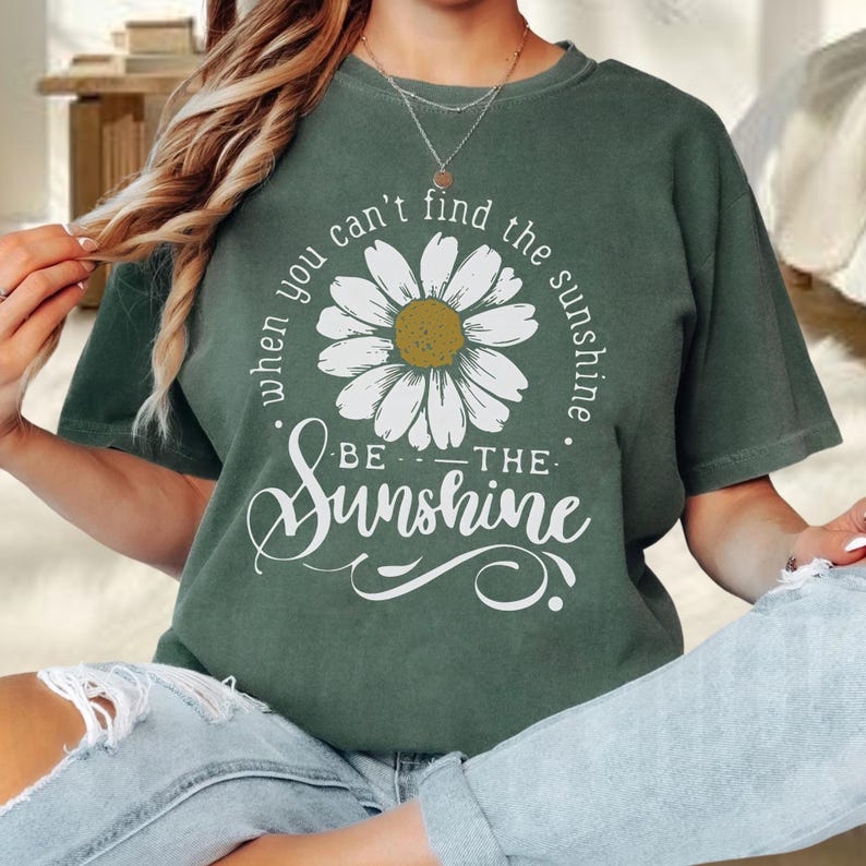 A woman wearing a green t-shirt with a white flower design and the text "When you can't find the sunshine, be the sunshine" printed on it.