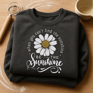 A black sweatshirt with a white daisy flower design and the text "When you can't find the sunshine, be the sunshine" printed on it, along with a wooden spoon and a small bowl.