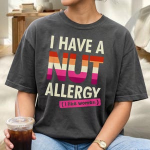 Puede incluir: Camiseta gris oscuro con el texto blanco "I HAVE A NUT ALLERGY" y un diseño "NUT" en colores arcoíris. Debajo, la palabra "ALLERGY" en blanco, y debajo, en un recuadro rosa, "(i like women)".