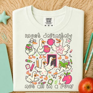 May include: A cream-colored t-shirt featuring a graphic print of white ducks with orange beaks and feet. The design includes school-themed items like books, pencils, an apple, and a chalkboard, with the text "most definitely not all in a row."