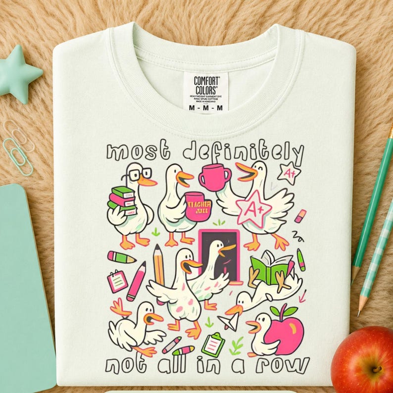 May include: A cream-colored t-shirt featuring a graphic print of white ducks with orange beaks and feet. The design includes school-themed items like books, pencils, an apple, and a chalkboard, with the text "most definitely not all in a row."