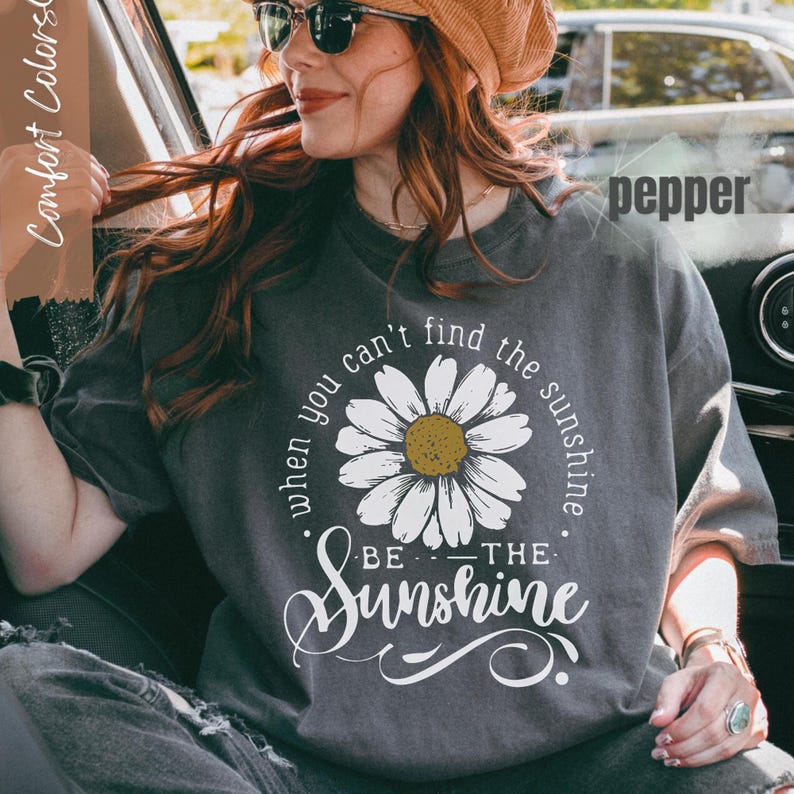 A woman wearing a gray t-shirt with a white flower design and the text "when you can't find the sunshine, be the sunshine" is sitting in a car.