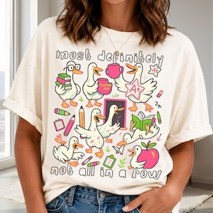 May include: An off-white t-shirt featuring a colorful graphic of cartoon ducks in school-themed activities. The text reads "most definitely" at the top and "not all in a row" at the bottom, surrounded by illustrations of books, pencils, and apples.