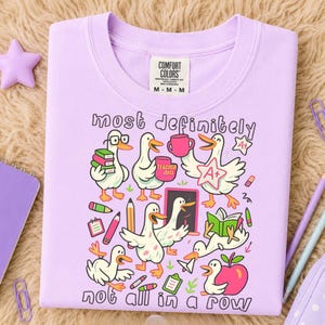 May include: A light purple t-shirt featuring a graphic print of several white ducks in various school-related activities. The text reads most definitely not all in a row. The design includes books, pencils, an apple, and an A+ star badge.