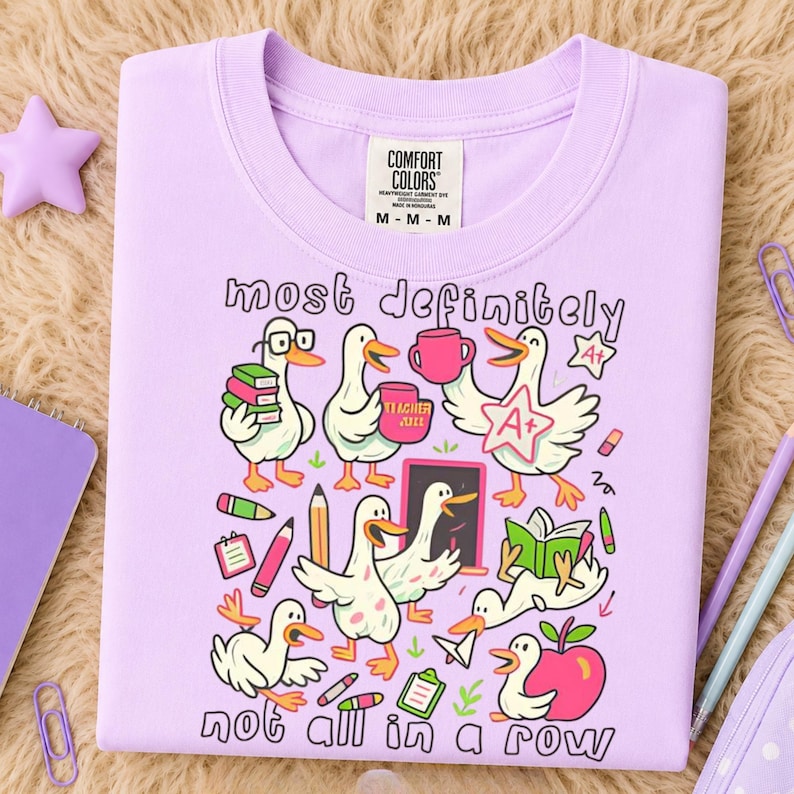 May include: A light purple t-shirt featuring a graphic print of several white ducks in various school-related activities. The text reads most definitely not all in a row. The design includes books, pencils, an apple, and an A+ star badge.
