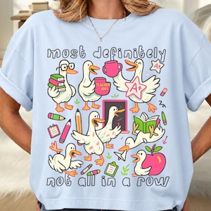 May include: A light blue t-shirt featuring a graphic print of white ducks engaged in school activities. The text reads "most definitely" at the top and "not all in a row" at the bottom. The ducks are shown with books, pencils, an apple, and an A+ star.