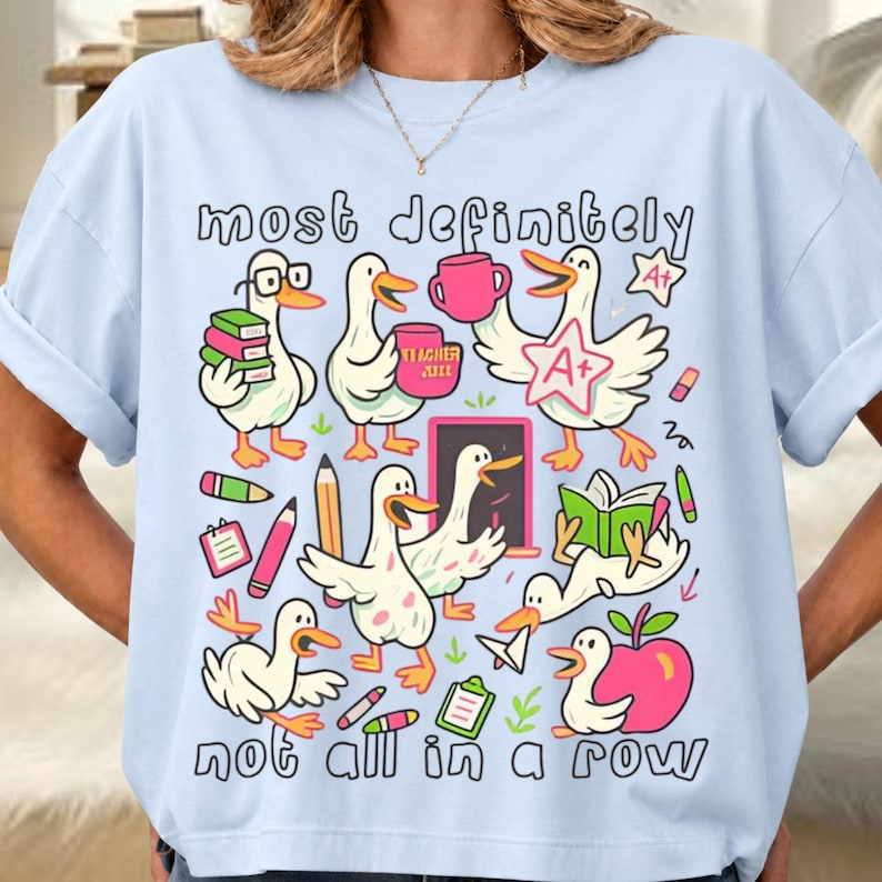 May include: A light blue t-shirt featuring a graphic print of white ducks engaged in school activities. The text reads "most definitely" at the top and "not all in a row" at the bottom. The ducks are shown with books, pencils, an apple, and an A+ star.