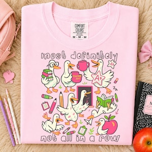 May include: A light pink t-shirt featuring a graphic print of white ducks in various school-themed activities. The text reads "most definitely" at the top and "not all in a row" at the bottom. The design includes books, pencils, and an apple.