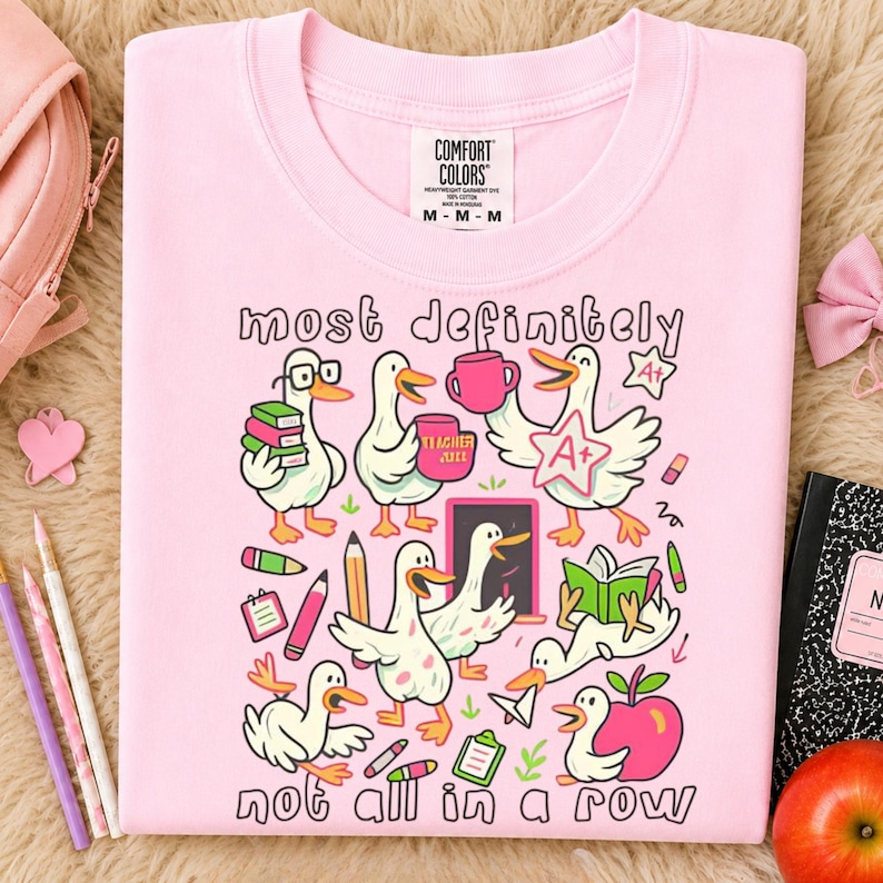 May include: A light pink t-shirt featuring a graphic print of white ducks in various school-themed activities. The text reads "most definitely" at the top and "not all in a row" at the bottom. The design includes books, pencils, and an apple.