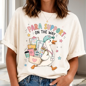 May include: An off-white t-shirt featuring a graphic of a white duck wearing a teal cap and a lanyard with a badge labeled Para. The duck carries a bag filled with a behavior chart, visual schedule, mini notes, and pencils. Text reads PARA SUPPORT ON THE WAY.