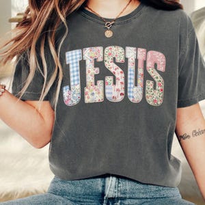 Puede incluir: Una camiseta de manga corta en color gris carbón con la palabra JESUS impresa en letras grandes. Cada letra está rellena con un patrón diferente, incluyendo cuadros vichy azules y blancos, estampados florales y rayas en tonos pastel.