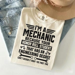 Könnte beinhalten: Ein cremefarbenes T-Shirt mit schwarzem Text: I'M A MECHANIC BECAUSE YOUR HONOR ROLL STUDENT THAT HAS AN ENGINEERING DEGREE CAN'T DESIGN IT RIGHT THE FIRST TIME. Das Design enthält grafische Symbole von Schrauben und einem Schraubenschlüssel.