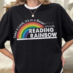 May include: A black t-shirt featuring a graphic with a rainbow arch and an open book icon. The text reads "Take a Look, it's in a Book!" and "READING RAINBOW" in white capital letters. The design is centered on the chest of the shirt.