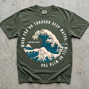 May include: An olive green short-sleeve t-shirt featuring a circular graphic of a large blue and cream-colored ocean wave. The text "WHEN YOU GO THROUGH DEEP WATES, I WILL BE WITH YOU" and "ISAIAH 43:2" is printed in white around the circular design.